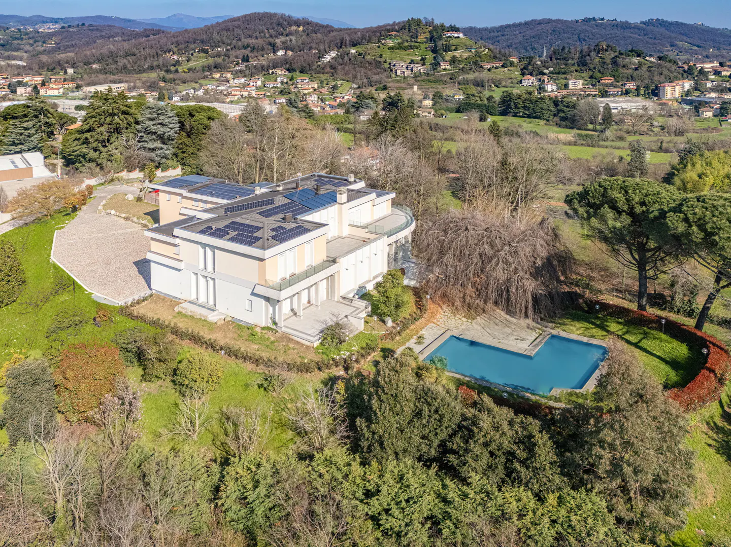 Aerial view of a modern, multi-story beige house with solar panels, a blue pool, and green trees against a mountain backdrop.