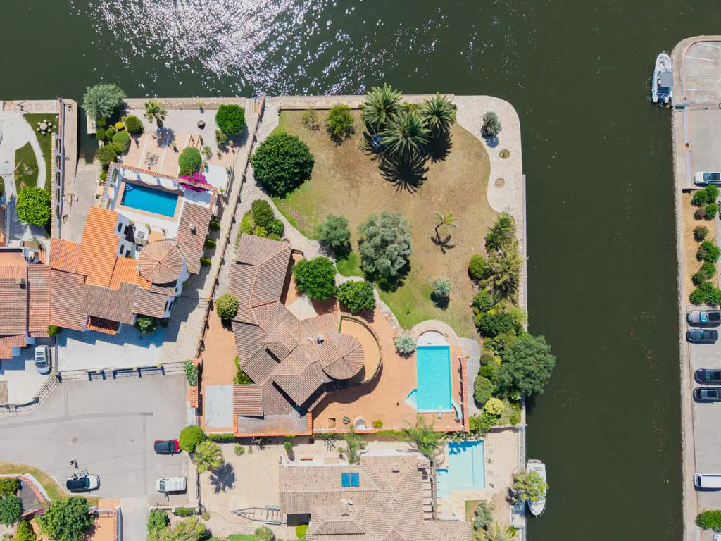 Aerial view of waterfront homes with terracotta roofs, swimming pools, lush gardens, and boats docked along the canal.