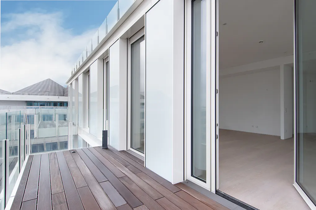 A modern apartment balcony with wood flooring, glass railings, and sliding glass doors leading to a bright, empty room.