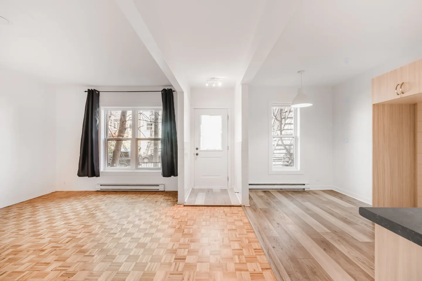 Bright, airy room with parquet floors, white walls, and black curtains framing a window. A white door leads to another room with wood floors.