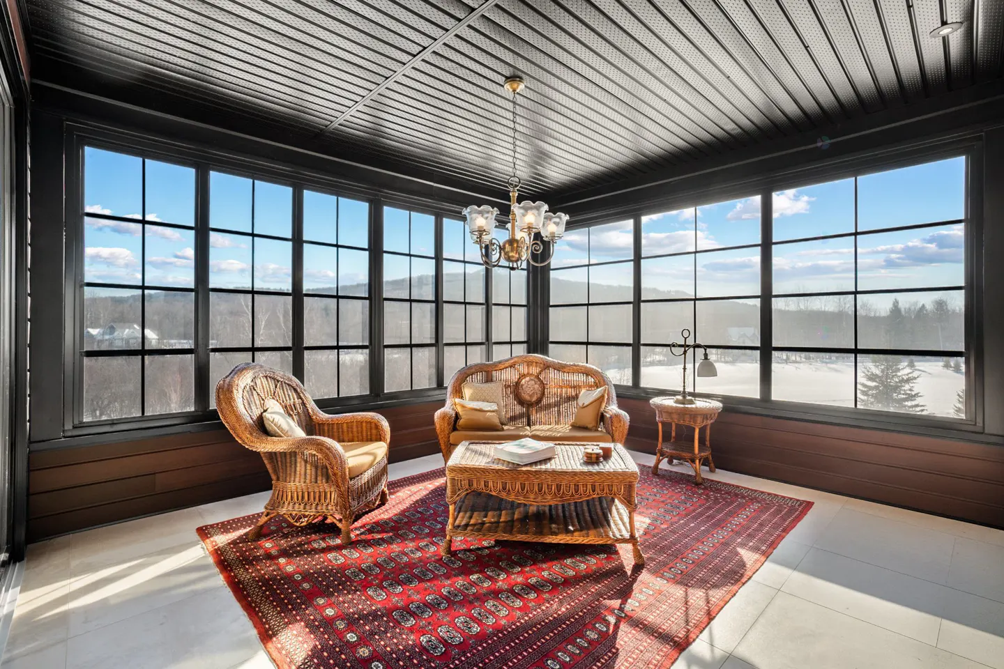 Sunroom with wicker furniture, red rug, and large windows overlooking a snowy landscape.