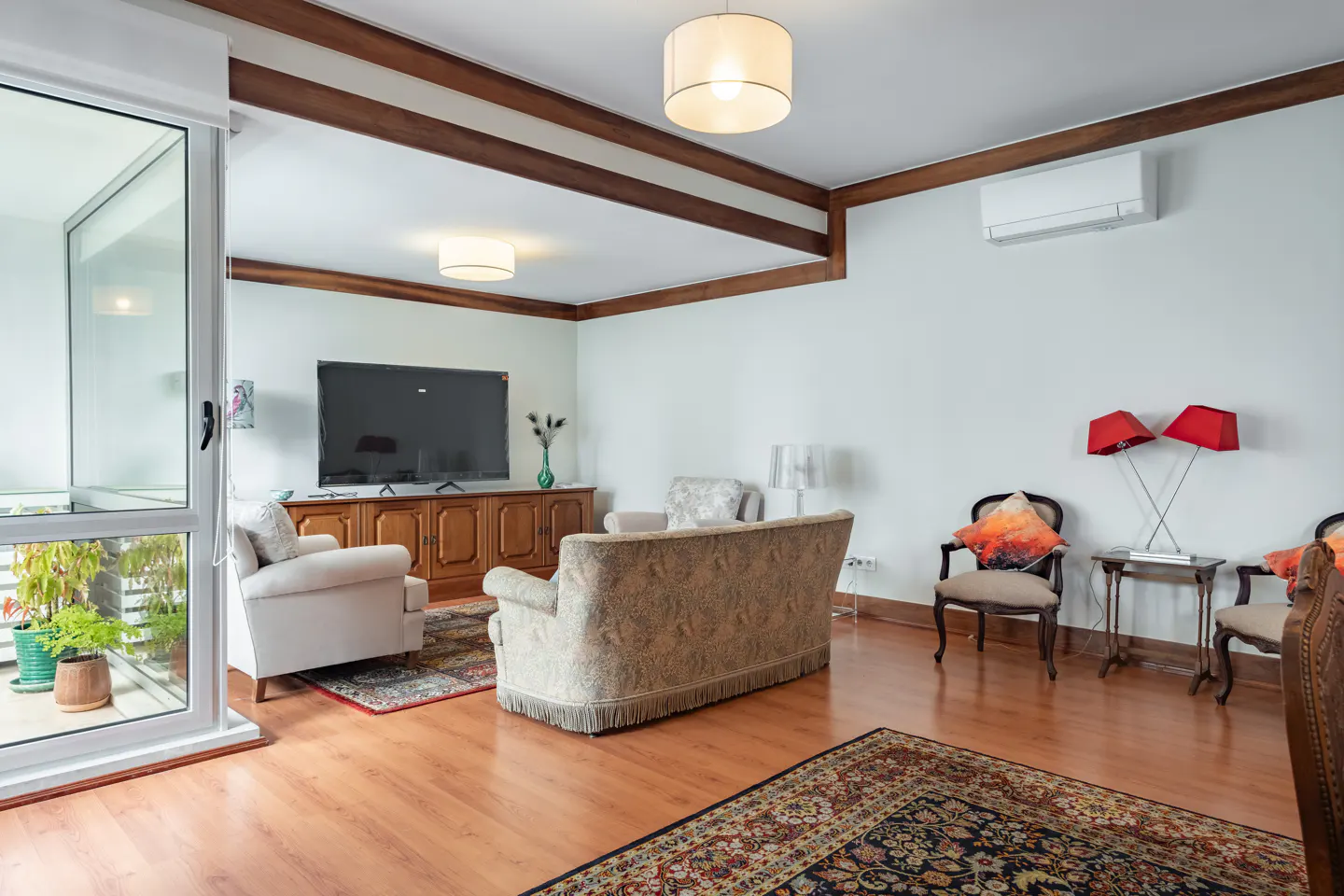 Living room with wood floors, white walls, and brown trim. A TV sits above a wooden cabinet. A balcony is visible through a sliding glass door.