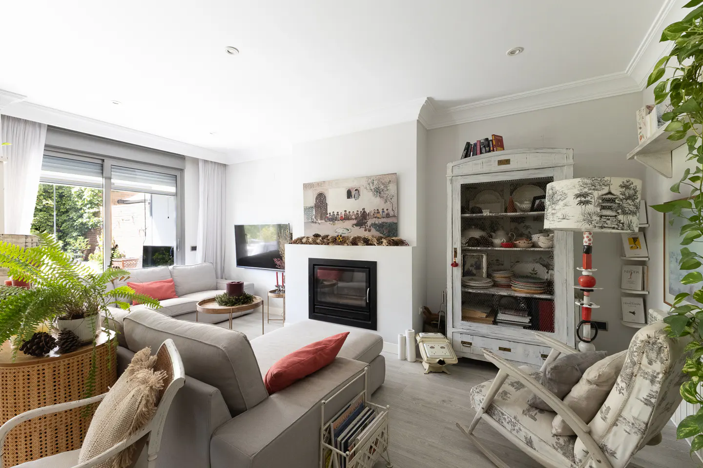 Bright living room with a fireplace, grey sofa, rocking chair, and a white vintage cabinet filled with decorative items.