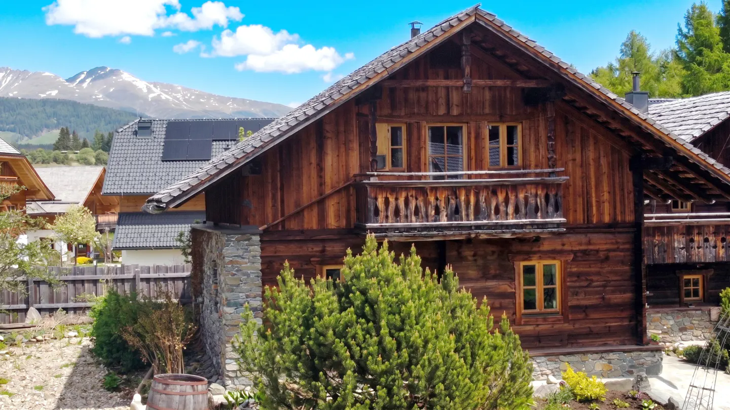 A two-story brown wooden chalet with a balcony, stone foundation, and mountains in the background.