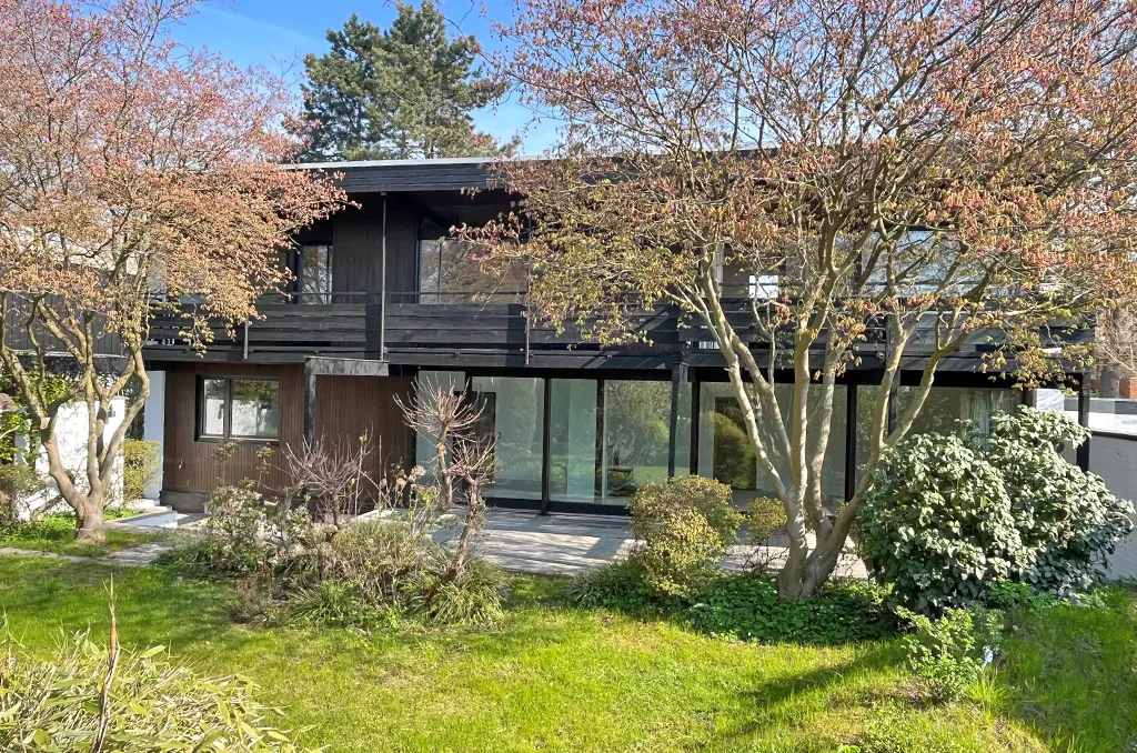 Two-story modern house with black trim, large windows, and a balcony, surrounded by trees and a green lawn.