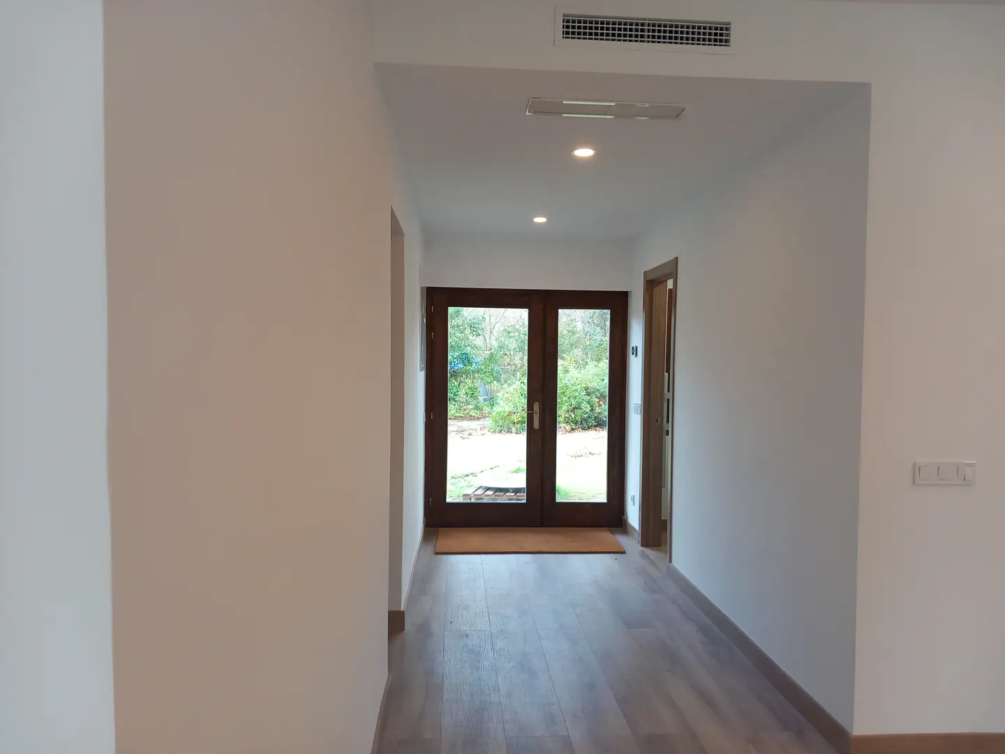 Bright hallway with light wood floors, white walls, and a dark wood double door leading to a green garden.