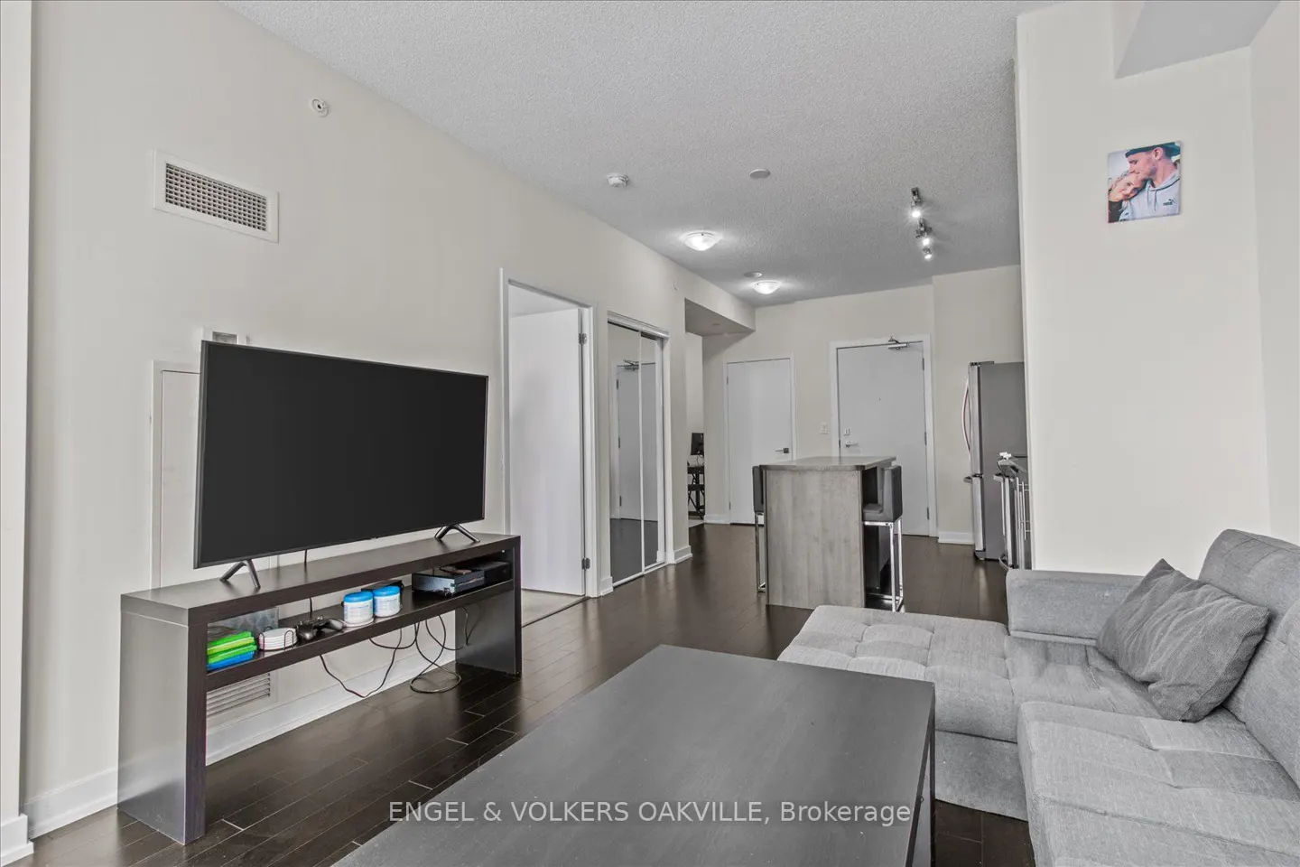 A modern living room with a gray sectional sofa, dark wood floors, and a large flat-screen TV. The walls are white, and there is a small kitchen area in the background.