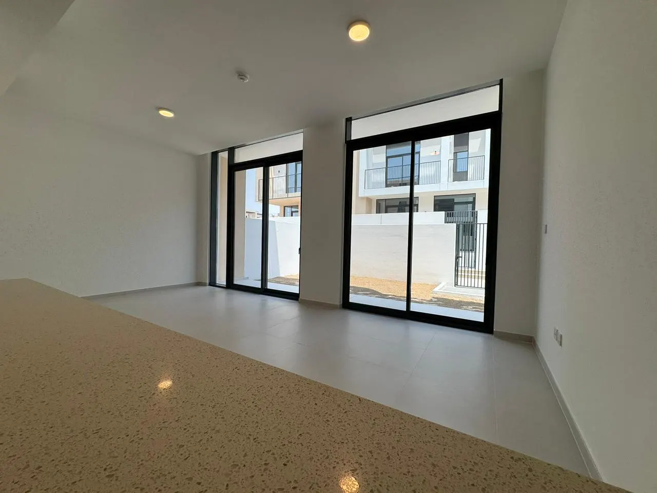 Bright, empty room with white walls and light tile floors. Large black-framed sliding glass doors lead to an outdoor area. A tan countertop is in the foreground.