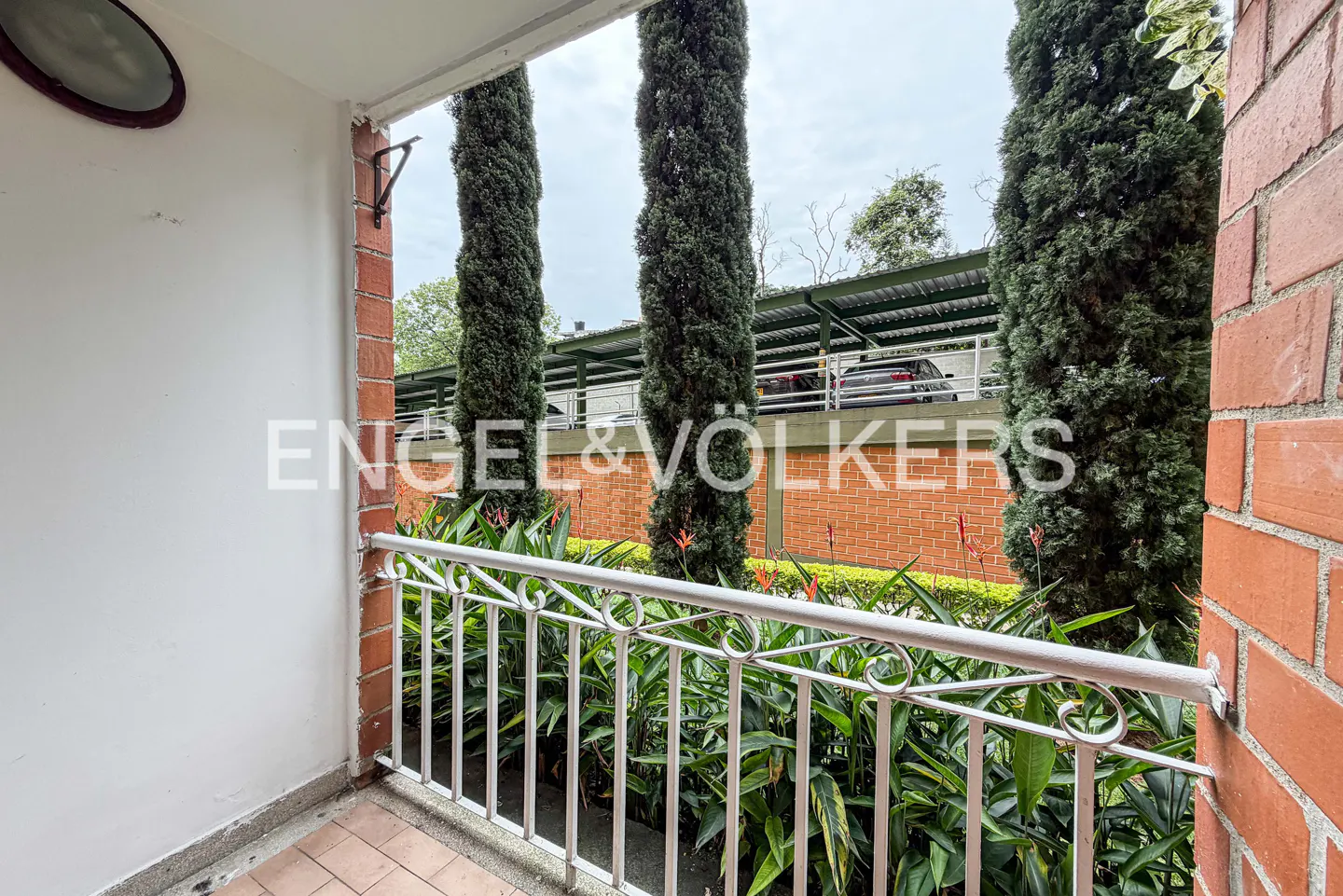 Balcony view with white railing, brick walls, and tall green trees. A parking structure is visible in the background.