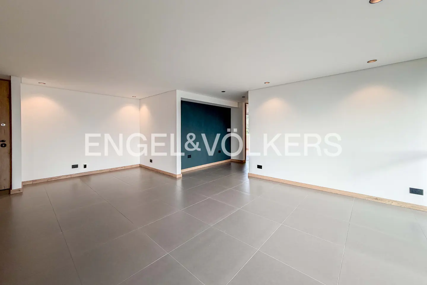 Empty room with gray tile floor, white walls, and a dark blue accent wall. Engel & Völkers logo is superimposed on the image.