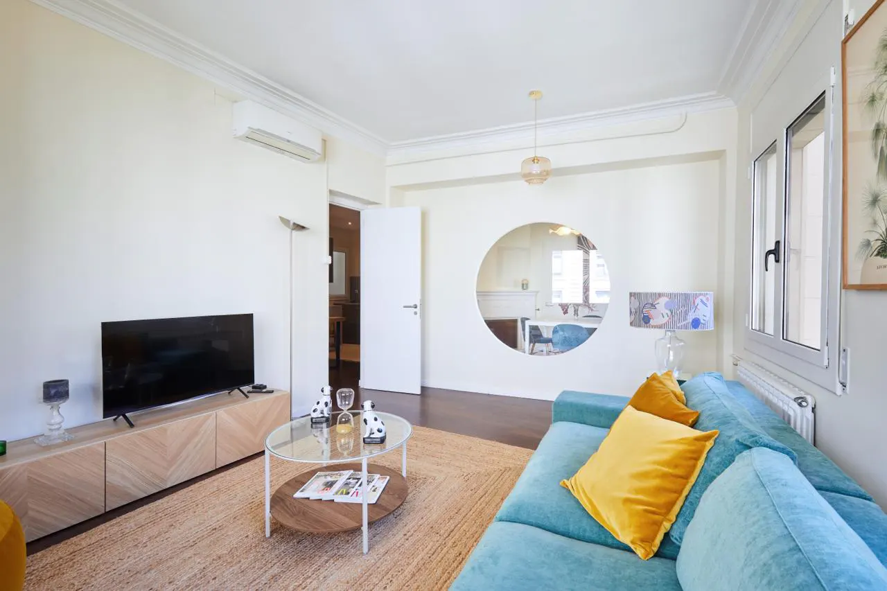 Bright living room with a blue sofa, yellow pillows, and a round mirror showing a dining area. A TV sits on a wooden cabinet.