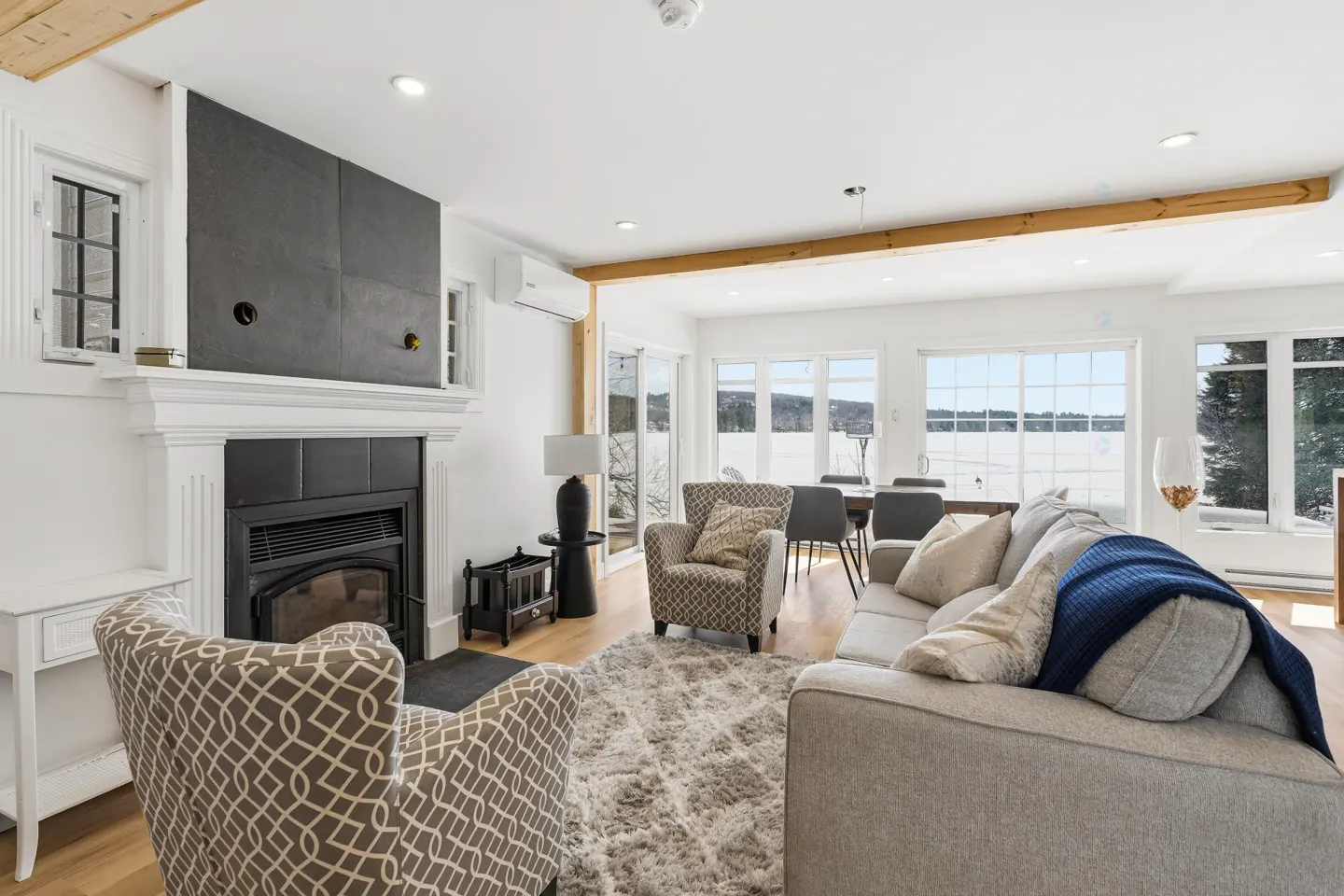 Bright living room with a fireplace, patterned armchairs, and a gray sofa with a blue blanket. Large windows overlook a snowy landscape.