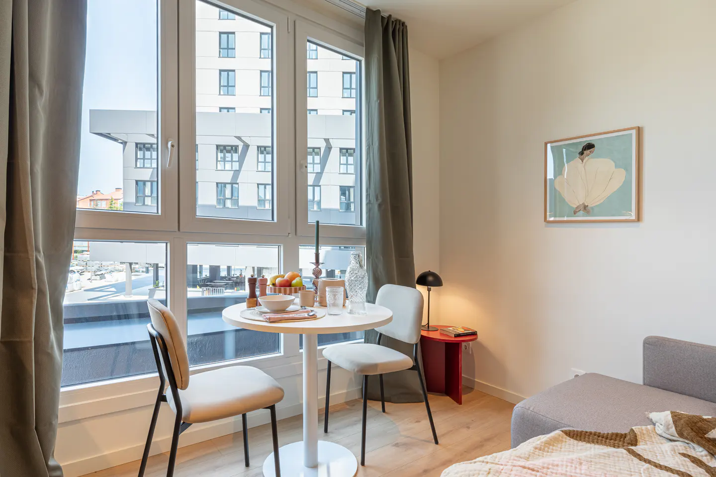 Bright apartment interior with a round white table set for two, chairs, and a large window with city views. A gray sofa and artwork add to the modern decor.