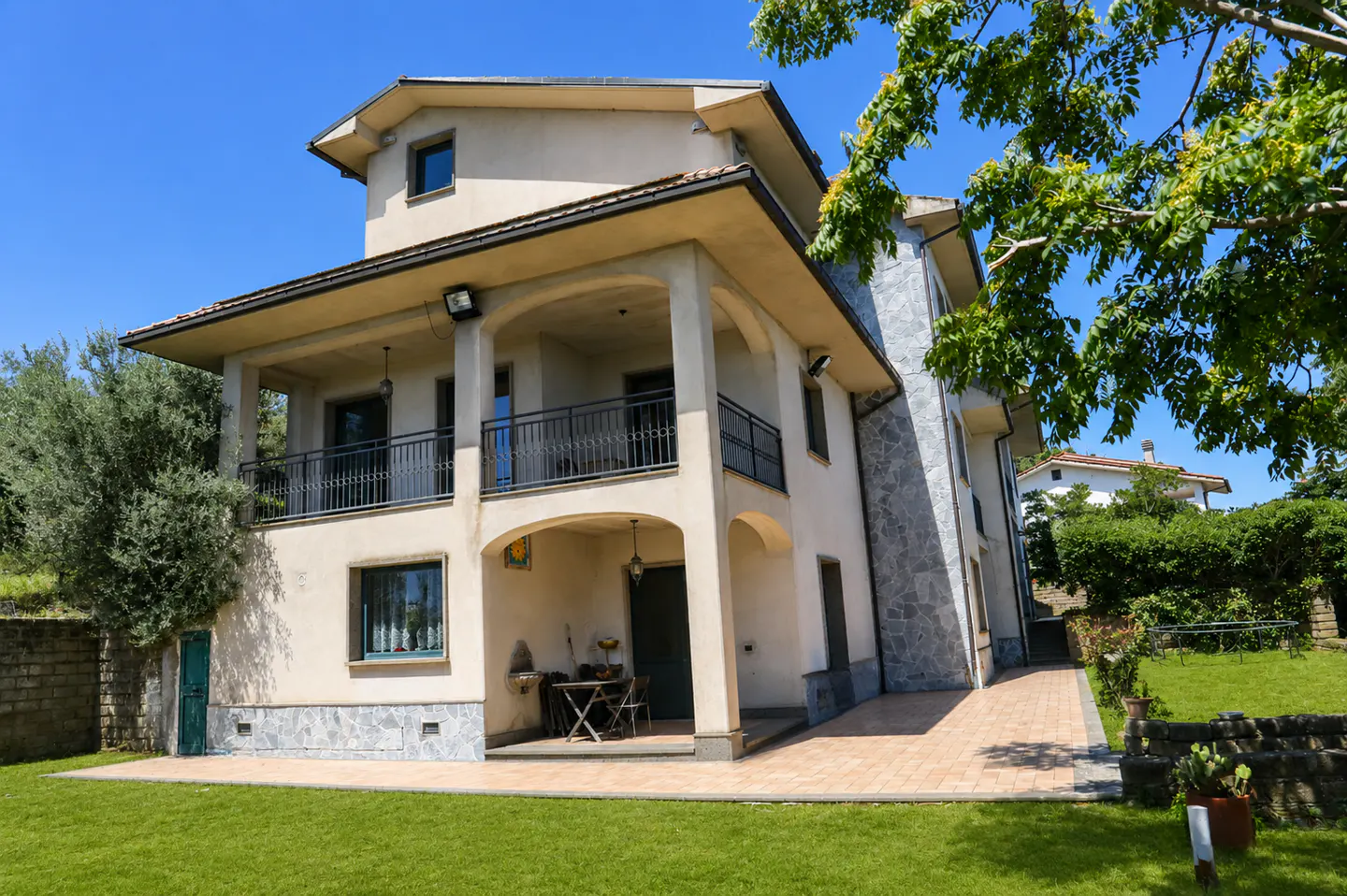 Two-story beige house with a balcony, black railings, and arched columns, surrounded by green lawn and trees under a blue sky.