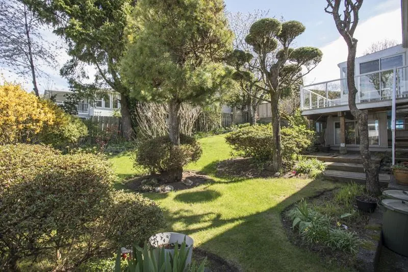 A lush green backyard with trees, bushes, and a house with a deck in the background.