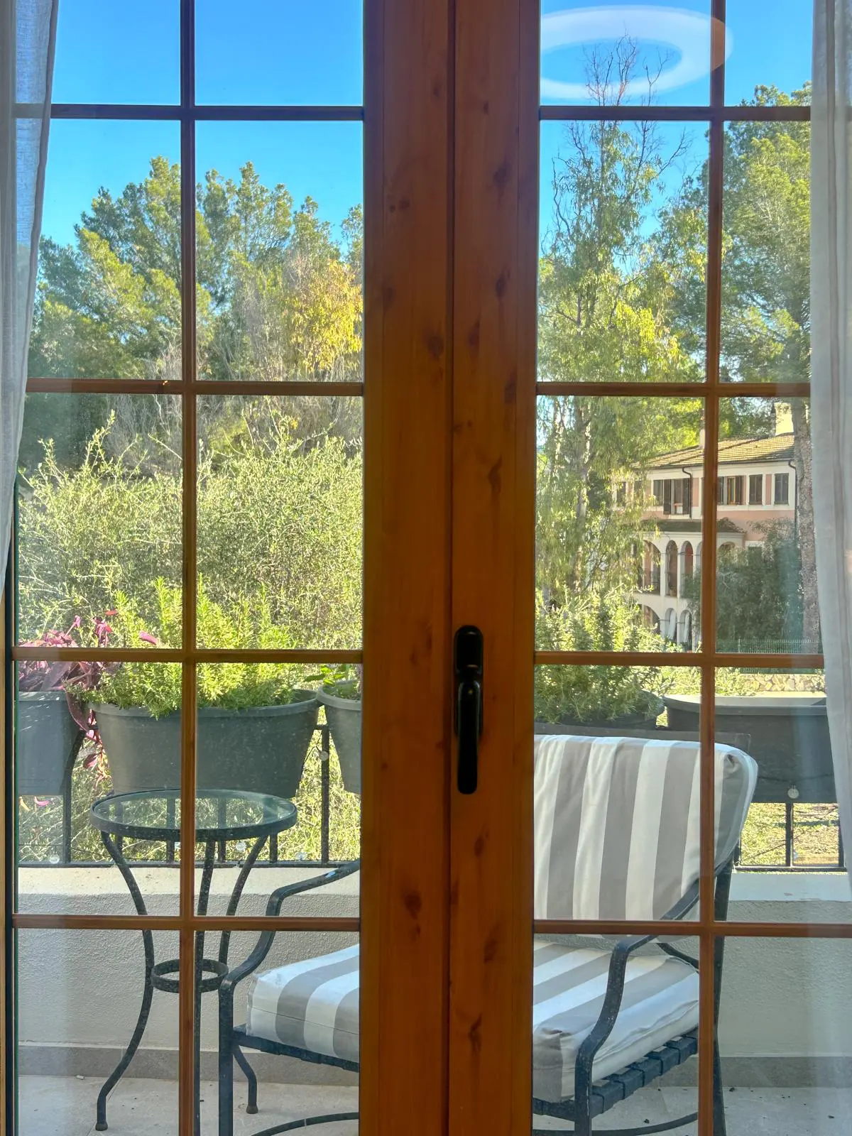 View through a window onto a patio with a striped chair, table, and potted plants, with a building and trees in the background.