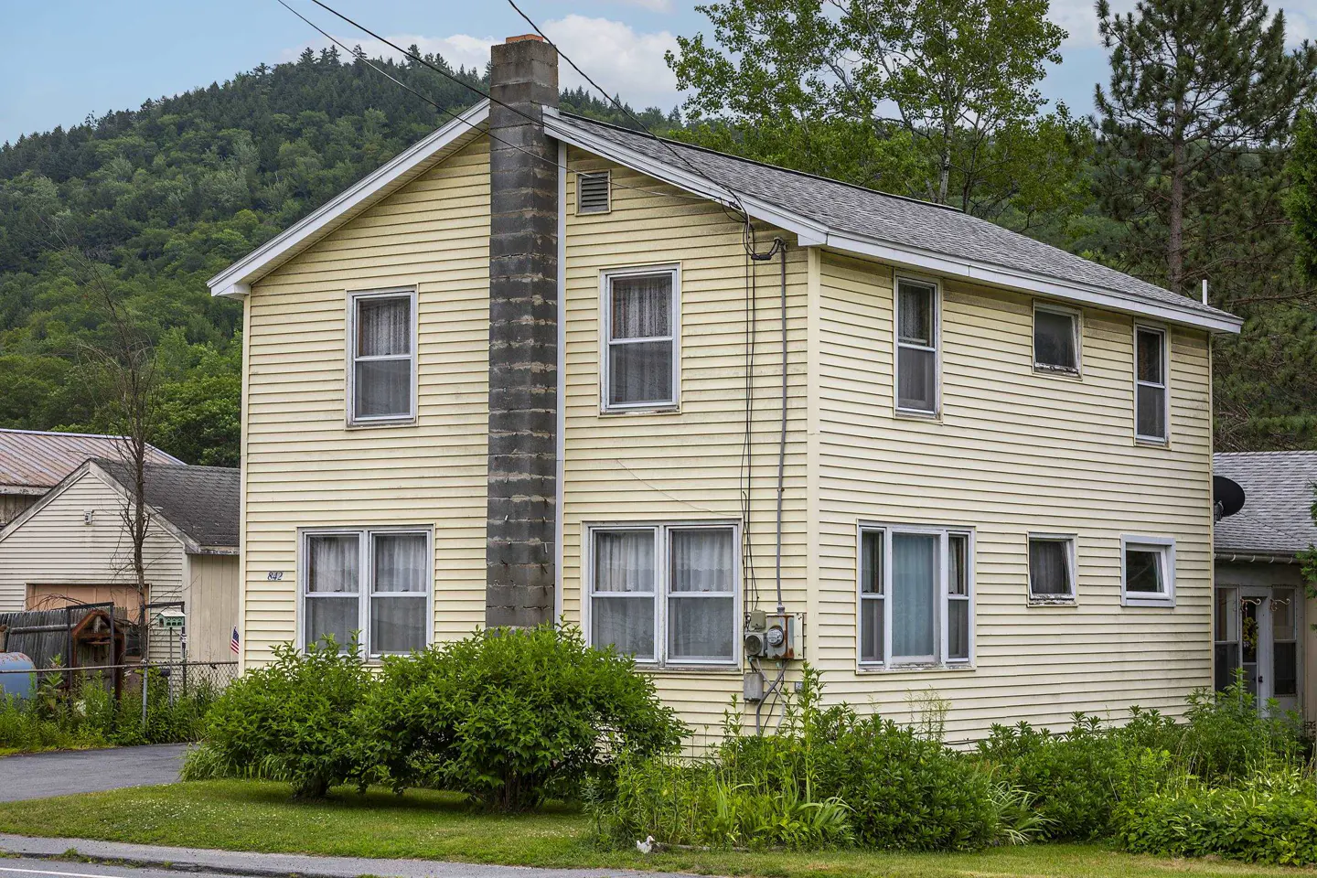 Two-story yellow house with a gray chimney and gray roof, surrounded by green bushes and trees.