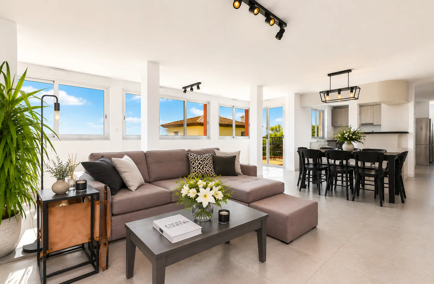 Bright, open-plan living space with a gray sectional sofa, coffee table, and dining area with black chairs. Large windows offer a view of a blue sky.