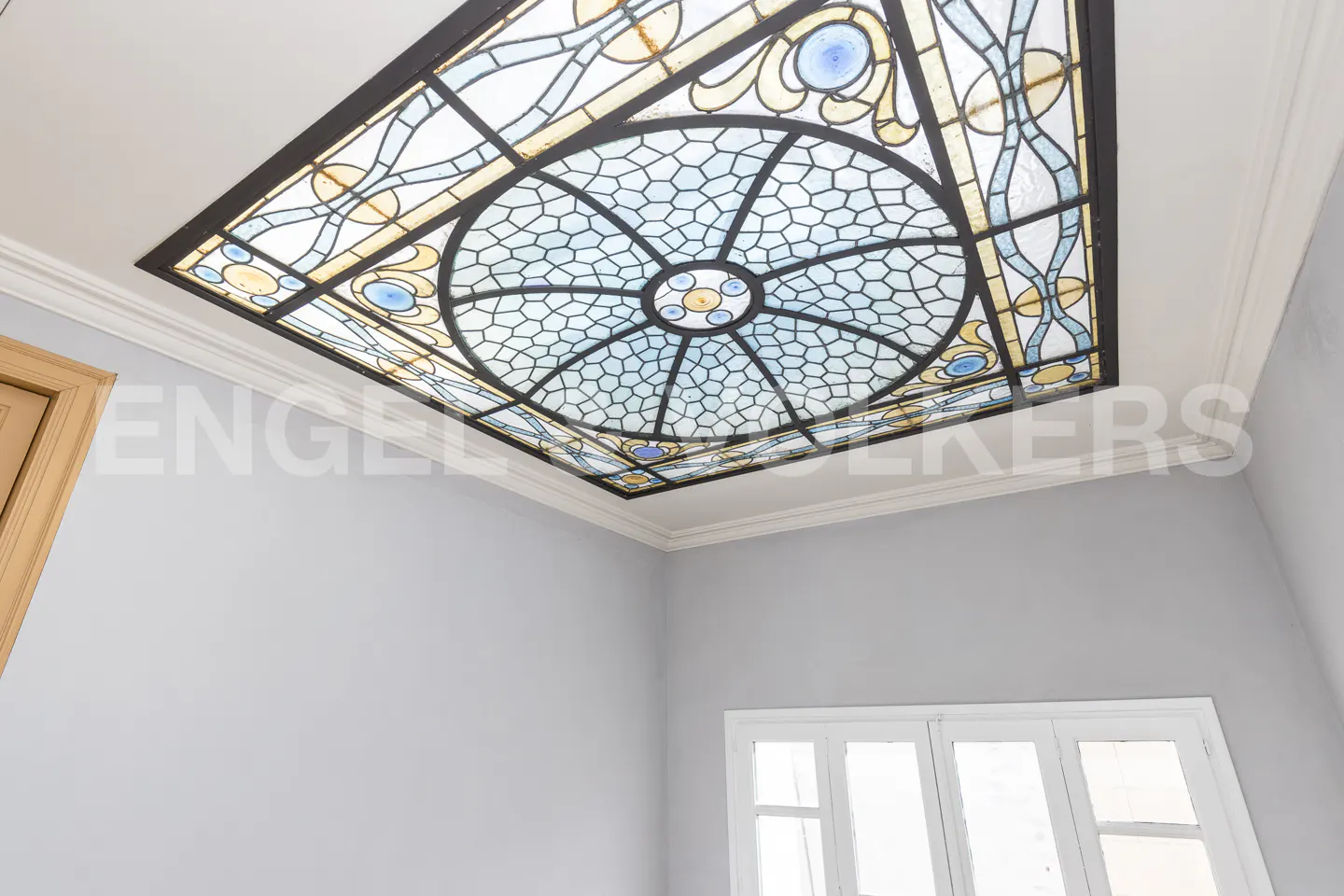 Looking up at a rectangular stained glass ceiling with blue, white, and yellow geometric patterns. Gray walls and a white window are visible.