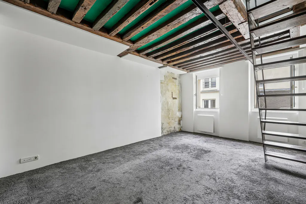 Empty room with grey carpet, white walls, exposed wooden ceiling beams with green accents, and a metal staircase.