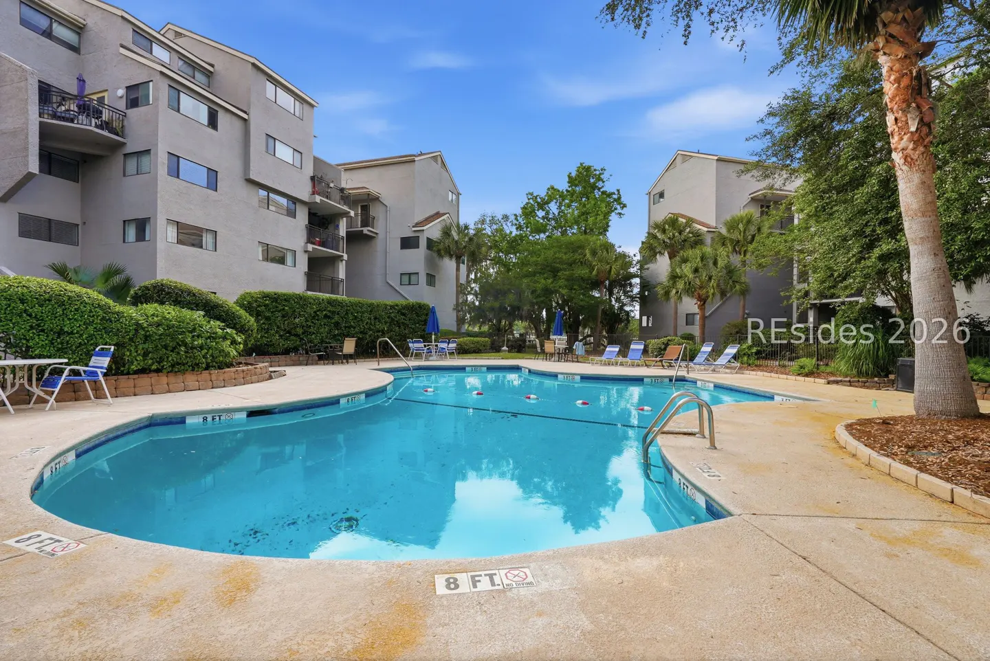 A bright blue swimming pool is surrounded by condos, trees, and lounge chairs on a sunny day.