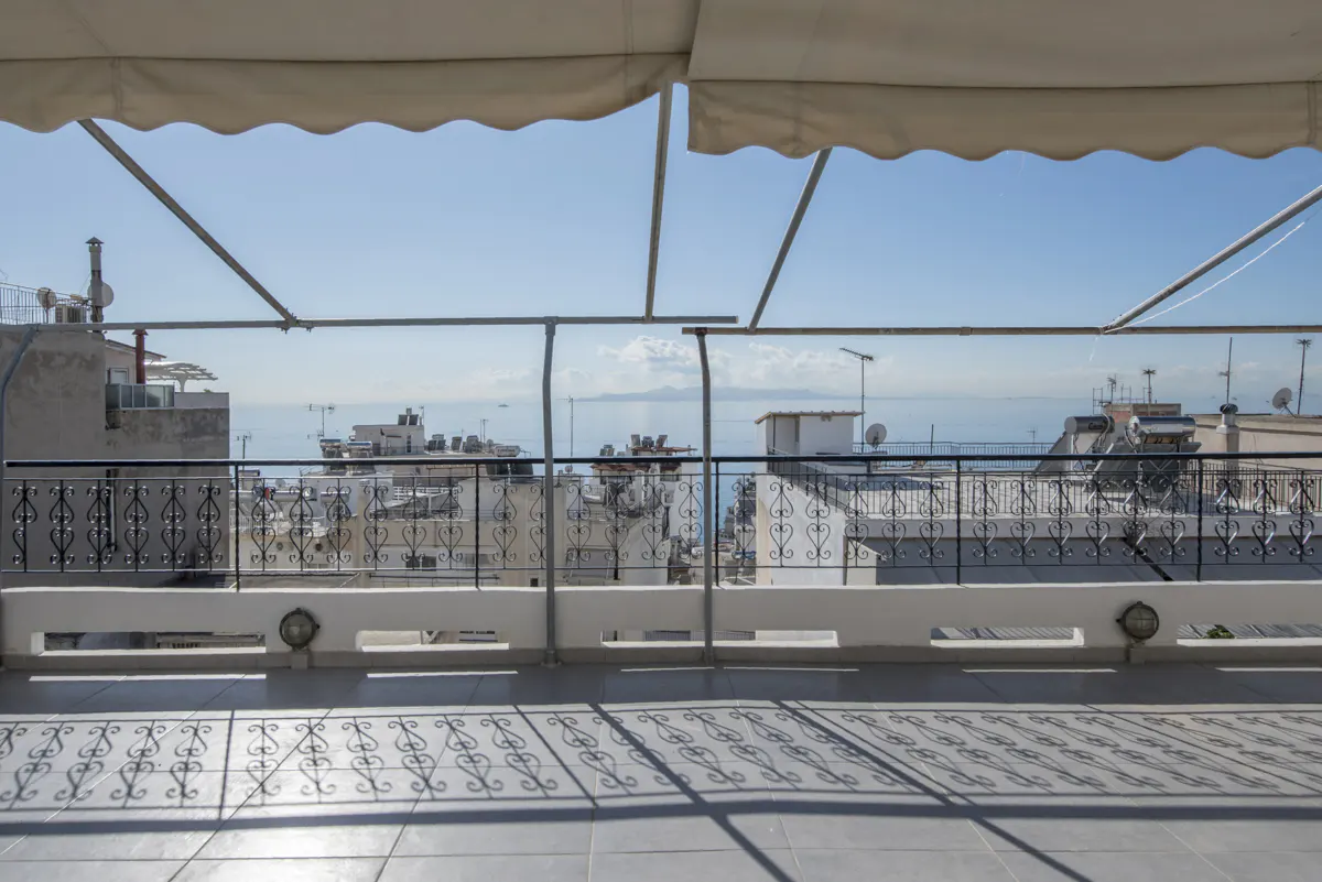 Balcony view of city buildings and blue sea under a white awning. Black wrought iron railing casts shadows on the gray tiled floor.