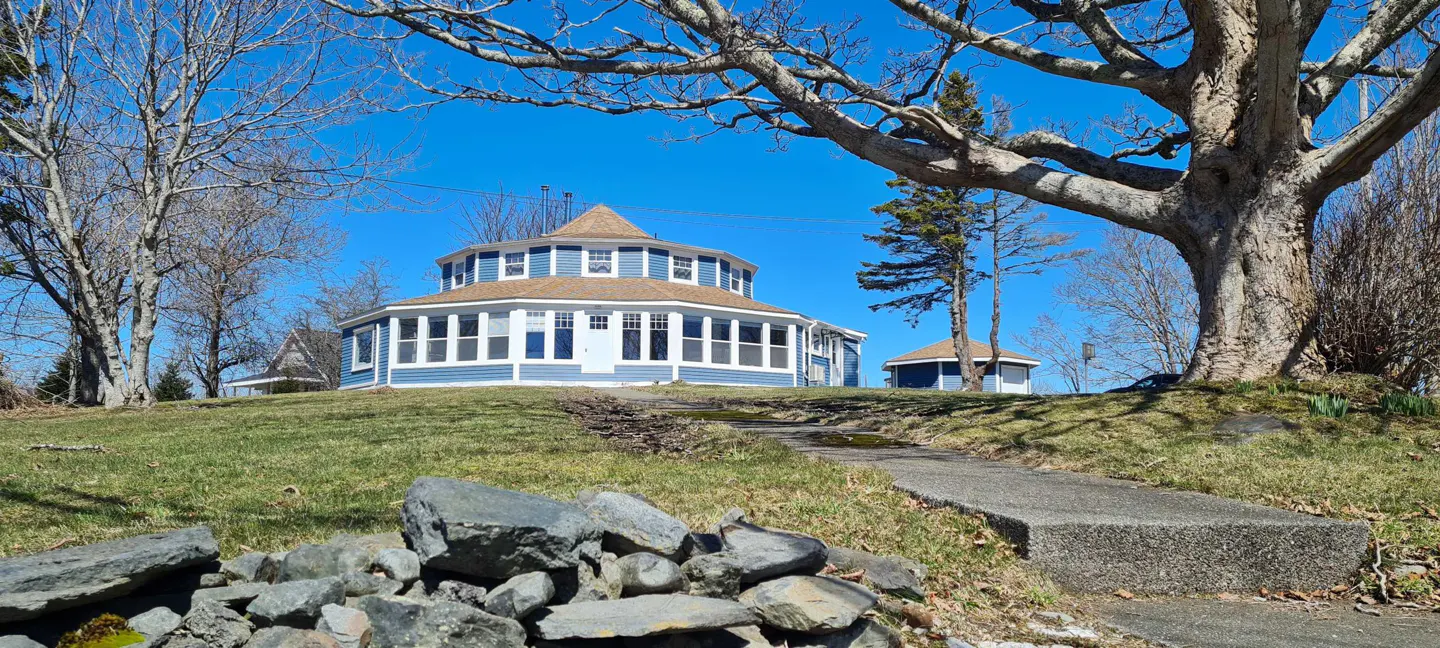 Blue, two-story, round house with a brown roof on a green lawn under a blue sky. A stone path leads to the house.