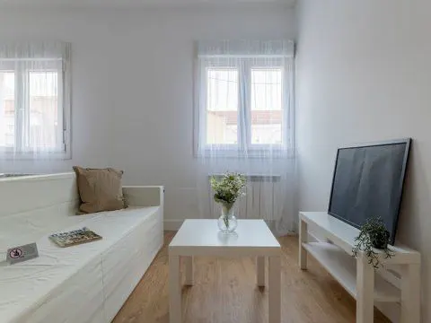 Bright living room with white walls, wood floors, and two windows with sheer curtains. A white sofa, table with flowers, and TV stand are also visible.