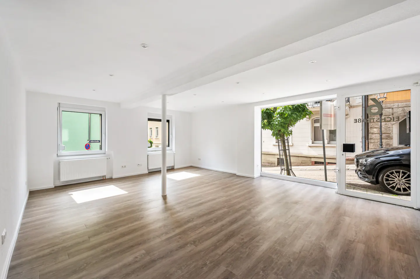 Bright, empty room with wood floors and white walls. Large window shows street view with a black car parked outside.