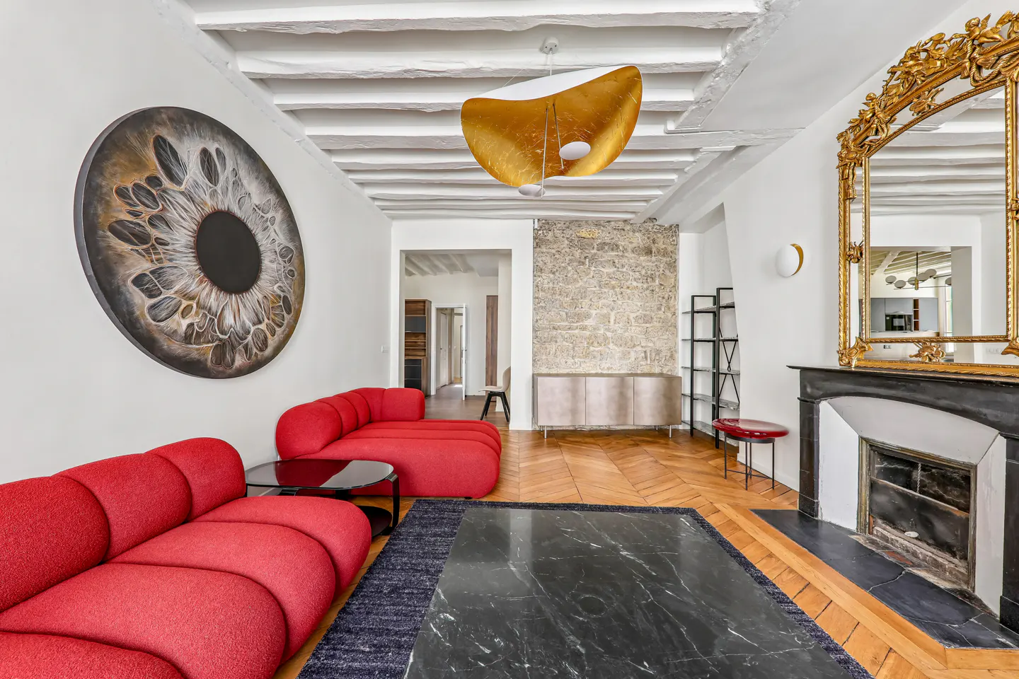 Living room with red sofas, black marble table, and a large eye art piece. Fireplace with gold mirror and exposed beams.