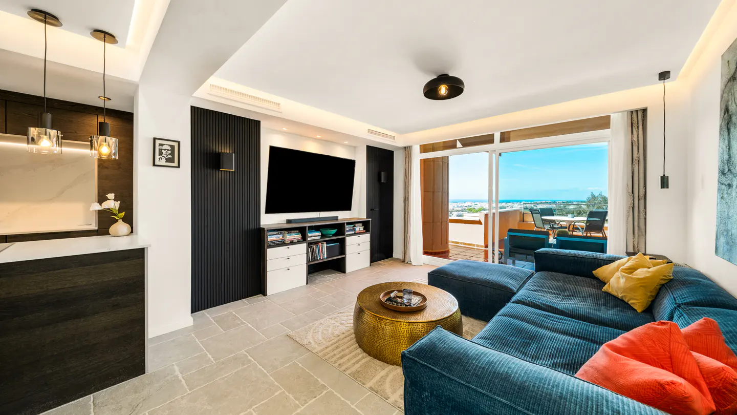 Modern living room with a blue sectional sofa, gold coffee table, and a TV. Balcony with a view of the ocean.