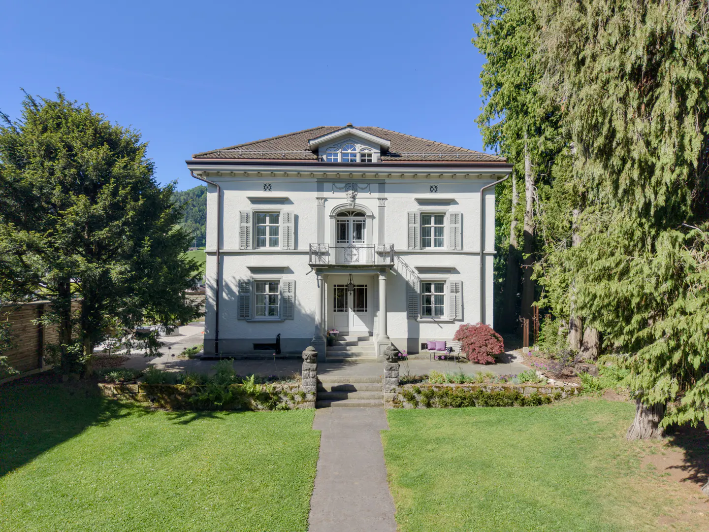 Two-story white house with gray shutters and a brown roof, surrounded by green trees and a lawn. A stone path leads to the front door.
