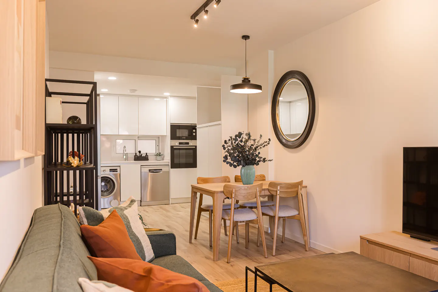 Open-concept living space with a green sofa, wood table with chairs, and a white kitchen in the background.