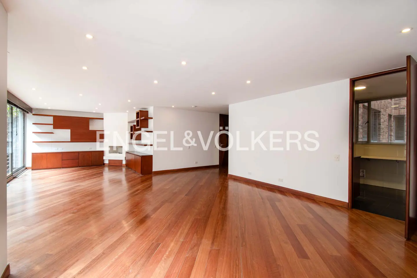 A spacious, bright living room with hardwood floors, white walls, and built-in wooden shelves and cabinets. A doorway leads to another room.