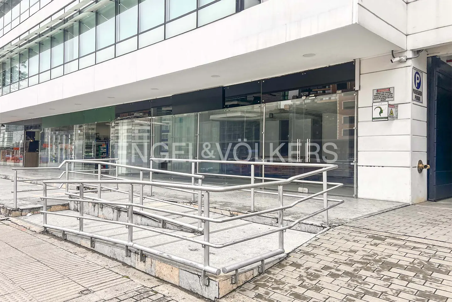 Exterior view of Engel & Völkers office with glass windows and a ramp with metal railings. Parking sign visible.