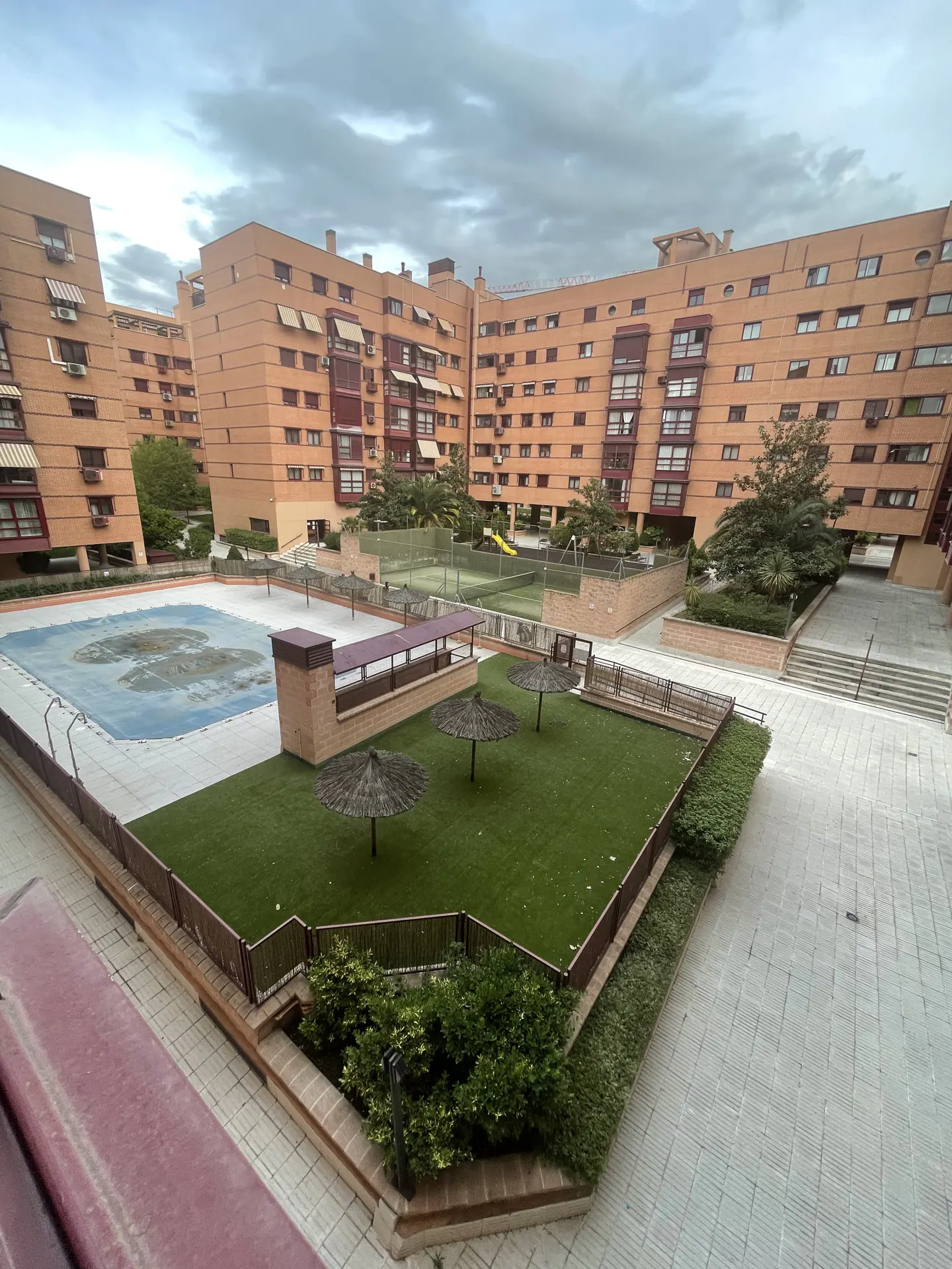 View of a brick apartment complex with a pool, tennis court, and green space. The sky is cloudy.