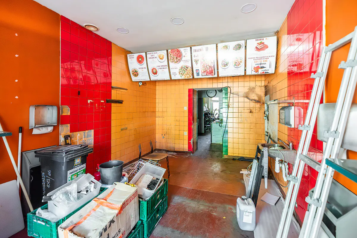 Interior of a vacant restaurant space with orange and red tiled walls. Menu boards hang above a doorway leading to a back room. Debris and equipment are scattered on the floor.