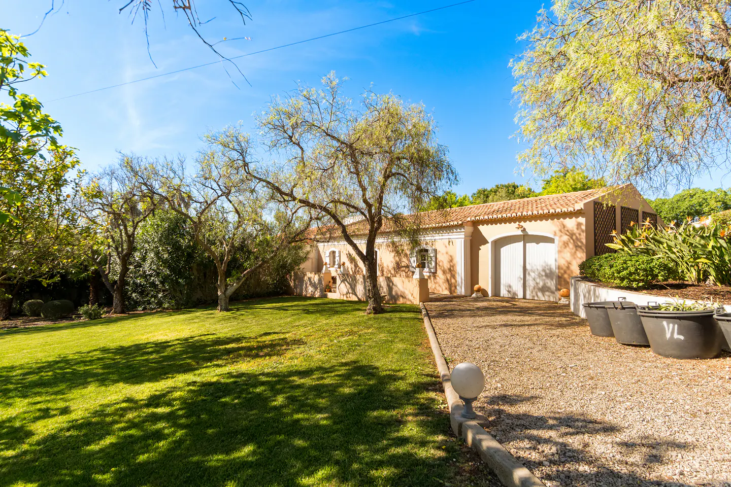 Exterior view of a tan house with a red tile roof, a green lawn, trees, and a gravel driveway on a sunny day.