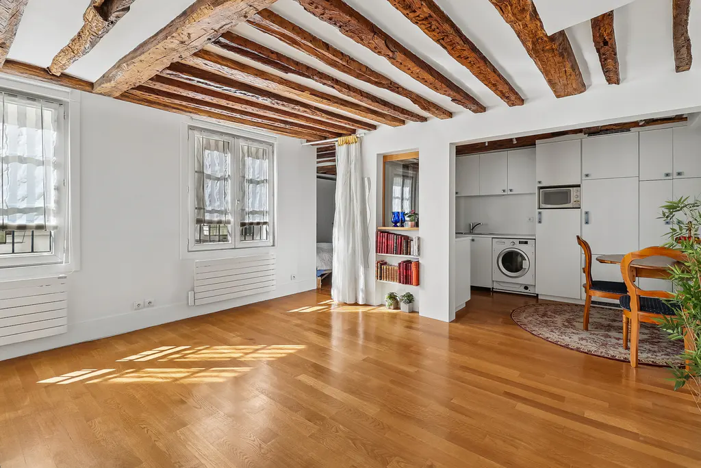 Bright apartment interior with wood floors, white walls, and exposed wood beams. Windows let in sunlight. A small kitchen is visible.