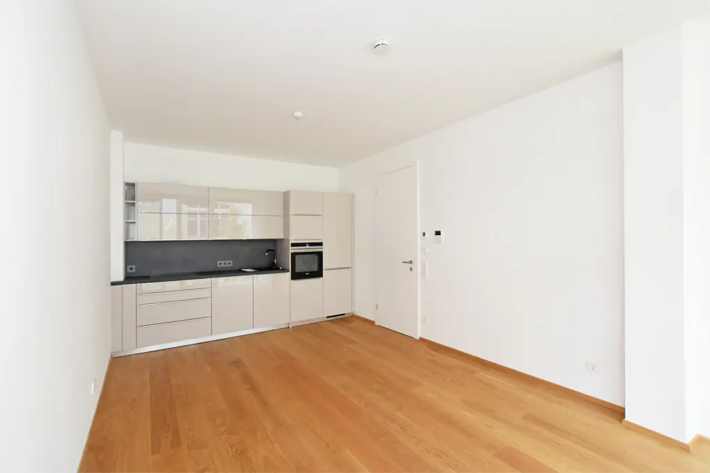 An empty apartment with light wood floors, white walls, and a modern kitchen with beige cabinets.
