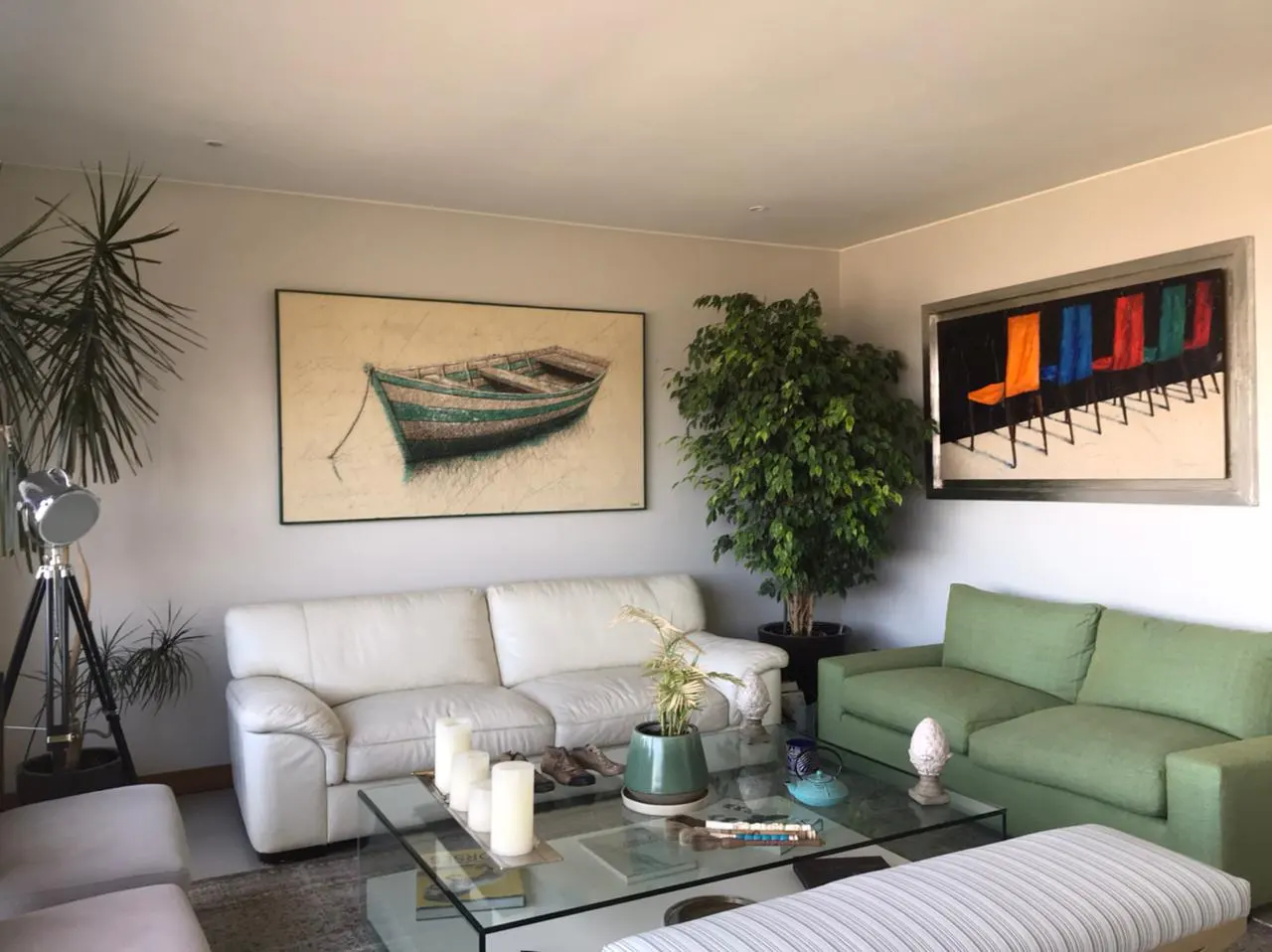 Living room with white and green sofas, glass coffee table, and artwork featuring a boat and colorful chairs.