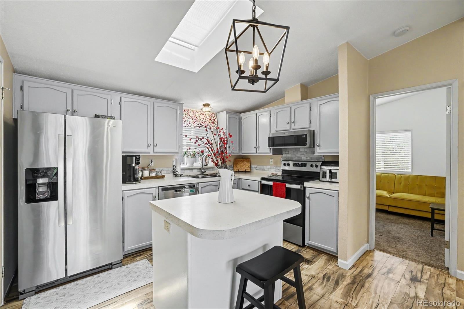 Modern kitchen with light gray cabinets, stainless steel appliances, a central island, and a view into a yellow living room. [AI-generated text]
