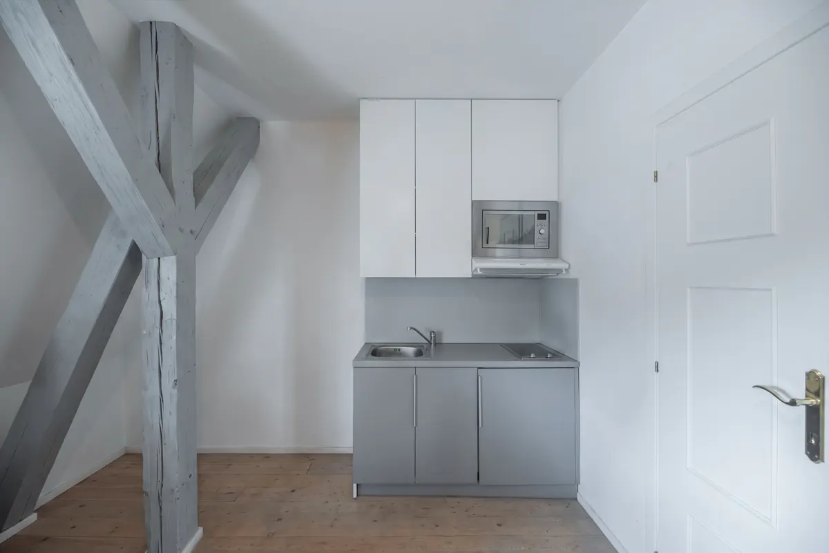 Small kitchen with grey cabinets, white upper cupboards, a microwave, and a sink, set against a white wall with wood beams.