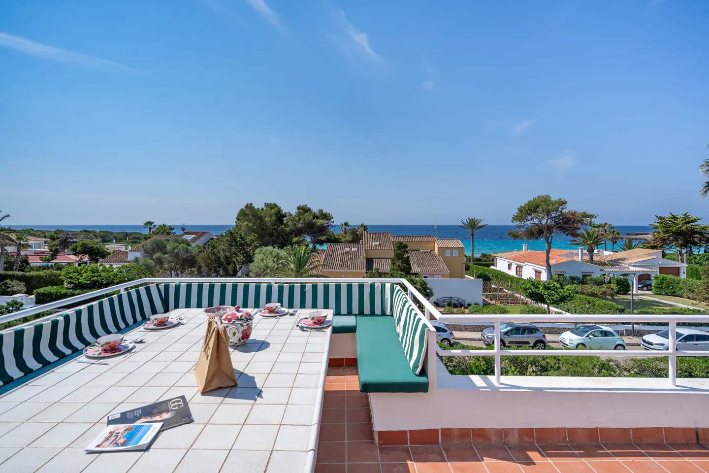 A sunny terrace with a tiled table, green-striped bench seating, and a scenic view of the Mediterranean sea and coastline.