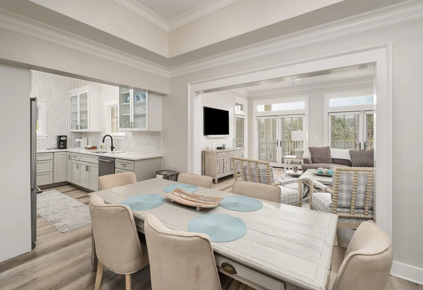 Open concept home with a dining table, kitchen, and living room. The dining table is white with beige chairs. The kitchen has gray cabinets and white backsplash. The living room has a gray sofa and chairs.