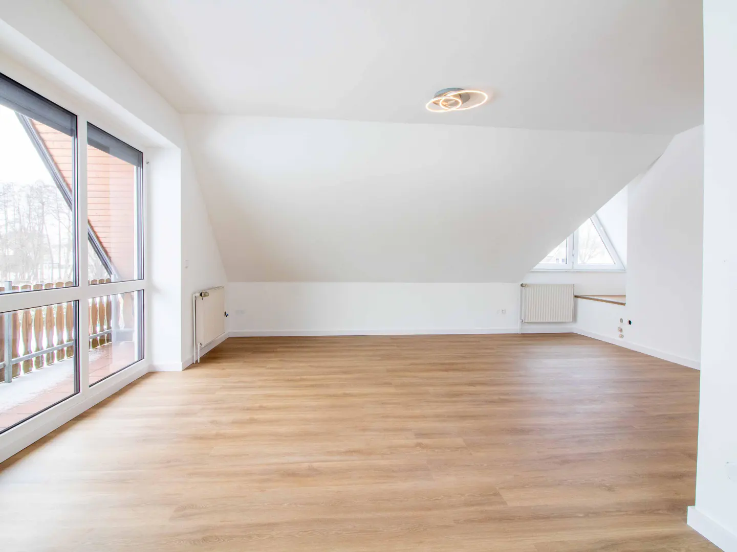 Bright, empty attic room with light wood floors, white walls, and large windows leading to a balcony. A modern light fixture is on the ceiling.