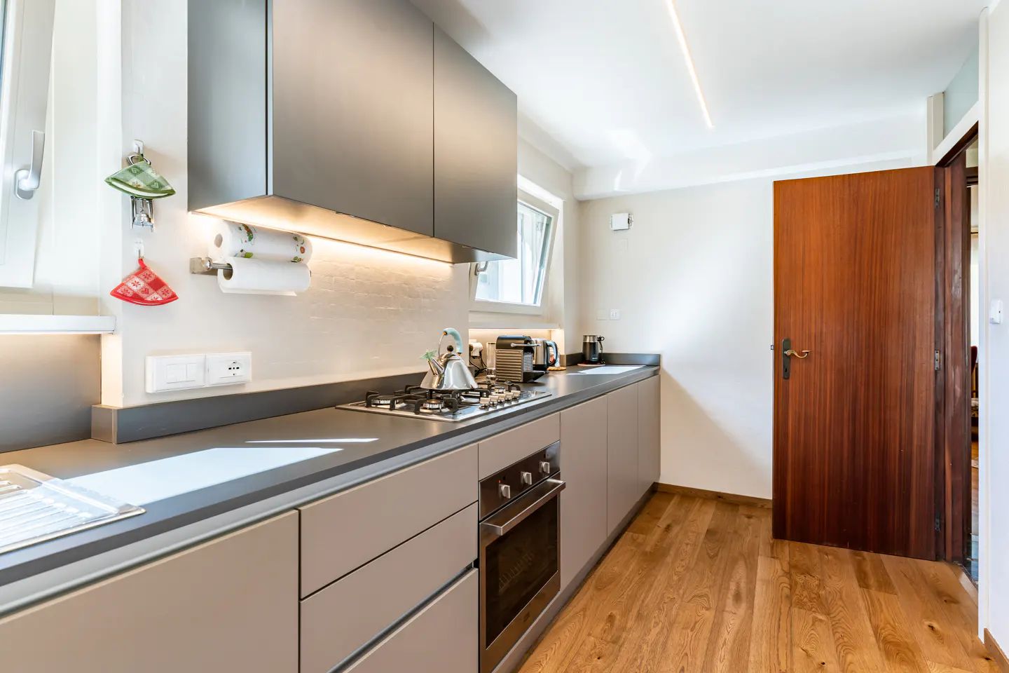 Modern kitchen with gray cabinets, stainless steel appliances, and wood floors. A kettle sits on the gas stove. A wooden door is ajar.