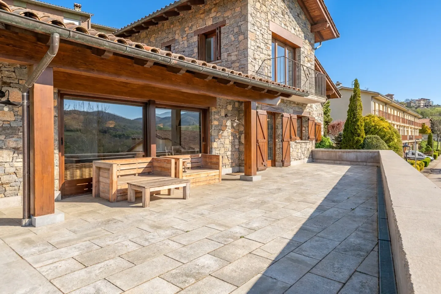 Stone house with a large patio featuring wooden furniture and shutters, under a clear blue sky.