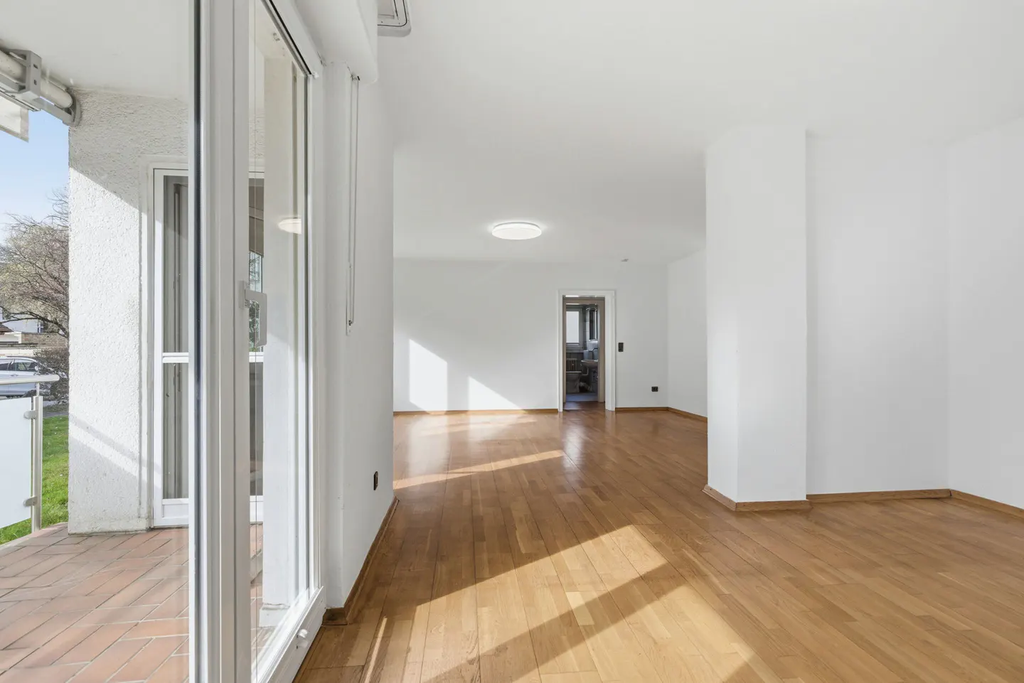 Bright, empty room with hardwood floors and white walls. Sliding glass doors lead to a balcony. A doorway leads to another room.