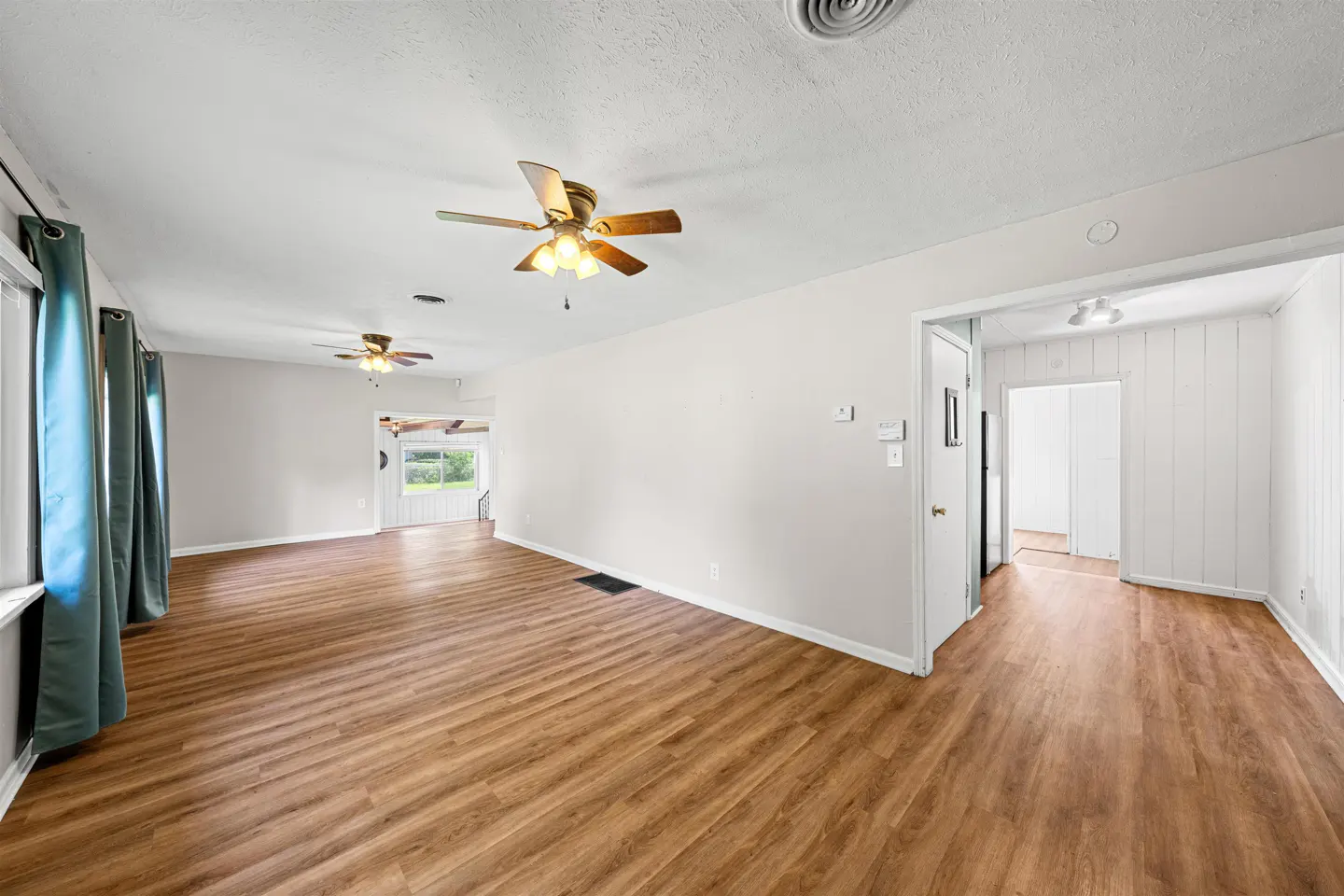 Spacious living room with light wood floors, white walls, two ceiling fans, and blue curtains over large windows.